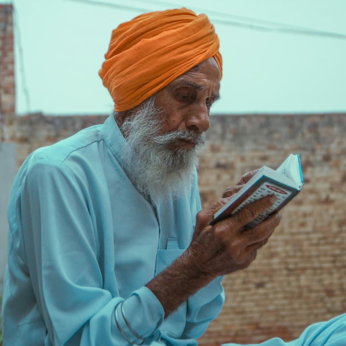 Mahinder Singh, an elderly man in an orange turban and pale blue kurta, sitting on a rooftop reading a small book.